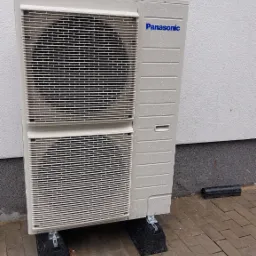 Zewnętrzna jednostka pompy ciepła Panasonic, zamontowana na czarnych podkładkach antywibracyjnych na kostce brukowej, widoczne krople wody po deszczu.