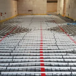 Białe rury ogrzewania podłogowego ułożone spiralnie na czarnej siatce z czerwonymi paskami w surowym wnętrzu budynku z bloczków.