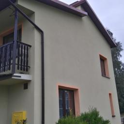 Firma Budowlana BRI Piotr Danek - Dwupiętrowy dom z balkonem, kremową elewacją i brązowymi obramowaniami okien. Widoczny żółty skrzynka gazowa, brązowa rynna i antena na dachu w tle.