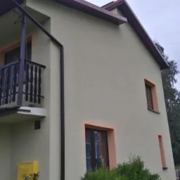 Dwupiętrowy dom z balkonem, kremową elewacją i brązowymi obramowaniami okien. Widoczny żółty skrzynka gazowa, brązowa rynna i antena na dachu w tle.