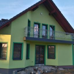 Firma Budowlana BRI Piotr Danek - Nowo otynkowany dom z balkonem i dekoracyjną balustradą, widoczny z rusztowaniem z boku, otoczony gruzem i zielenią.