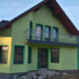 Nowo otynkowany dom z balkonem i dekoracyjną balustradą, widoczny z rusztowaniem z boku, otoczony gruzem i zielenią.