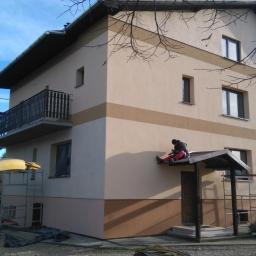 Firma Budowlana BRI Piotr Danek - Dwukondygnacyjny dom z balkonem po ociepleniu, z elementami dekoracyjnymi w odcieniach brązu. Pracownik na daszku wejścia, rusztowania, firmowy bus z reklamą.