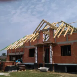 Budowa domu murowanego w trakcie realizacji: ściany z czerwonej cegły, drewniana konstrukcja dachu, widoczny pracownik na rusztowaniu. Etap surowy otwarty.