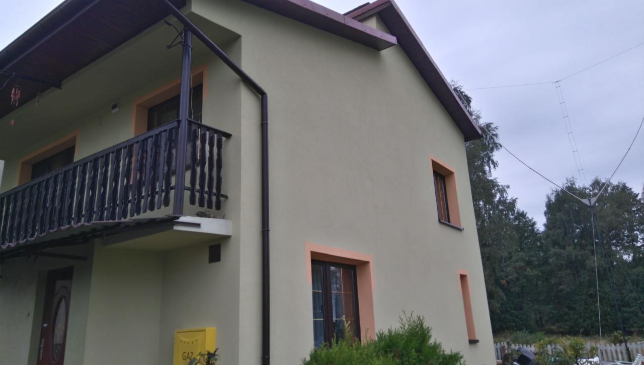 Dwupiętrowy dom z balkonem, kremową elewacją i brązowymi obramowaniami okien. Widoczny żółty skrzynka gazowa, brązowa rynna i antena na dachu w tle.