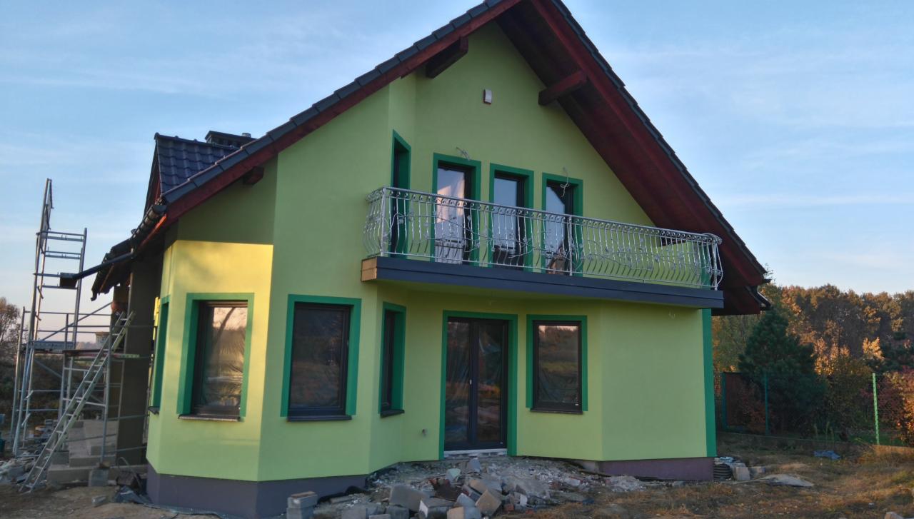 Nowo otynkowany dom z balkonem i dekoracyjną balustradą, widoczny z rusztowaniem z boku, otoczony gruzem i zielenią.