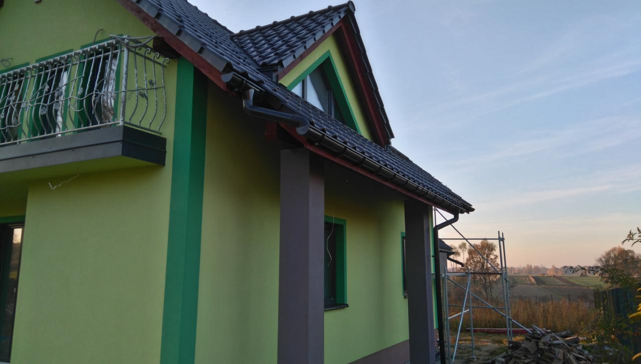 Żółto-zielony dom z czarnym dachem i balkonem z kutą balustradą. Widoczne rusztowania, pole w tle i fragmenty składowanych materiałów budowlanych.