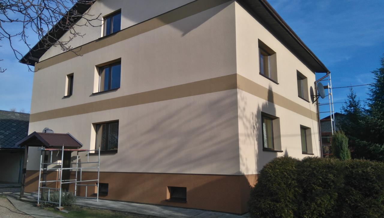 Dwukondygnacyjny dom z oknami i elewacją w odcieniach beżu i brązu, z widocznymi rusztowaniami przy wejściu i anteną satelitarną na bocznej ścianie.