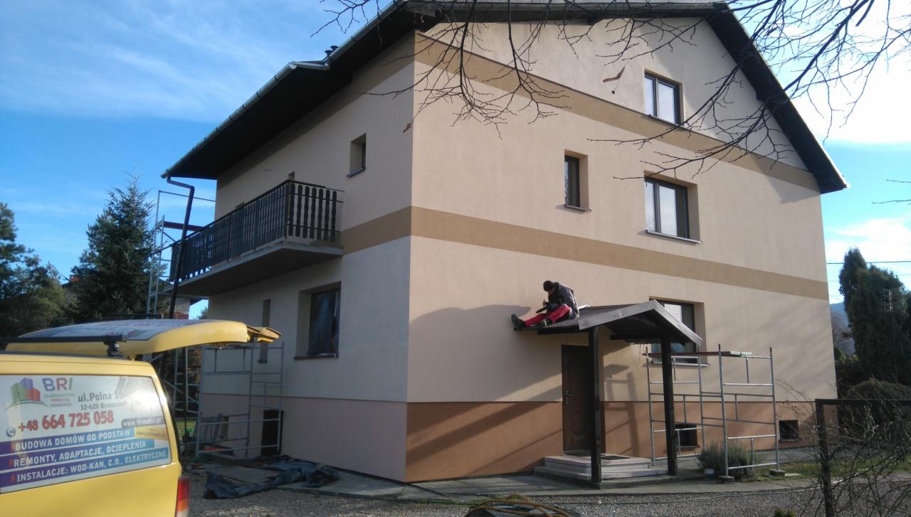 Dwukondygnacyjny dom z balkonem po ociepleniu, z elementami dekoracyjnymi w odcieniach brązu. Pracownik na daszku wejścia, rusztowania, firmowy bus z reklamą.