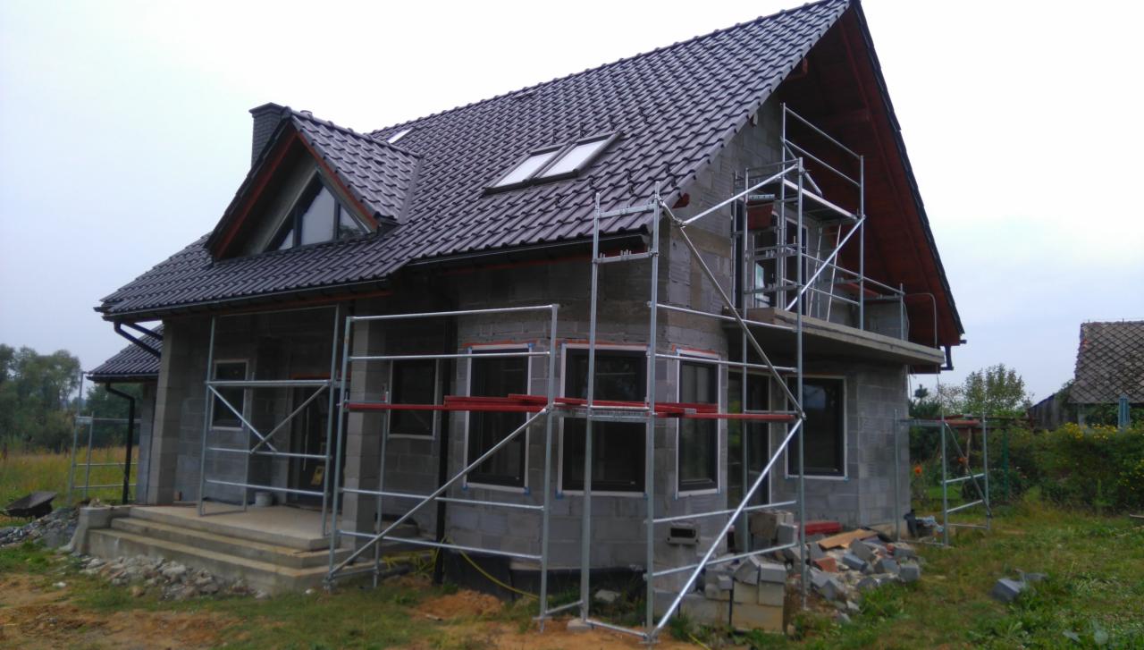 Dom w trakcie budowy z szarego bloczka, z ciemnym dachem i oknami. Widoczne rusztowania i elementy konstrukcyjne. Etap budowy przed ociepleniem i wykończeniem elewacji.