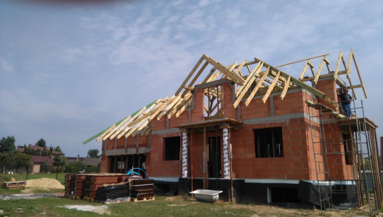 Budowa domu murowanego w trakcie realizacji: ściany z czerwonej cegły, drewniana konstrukcja dachu, widoczny pracownik na rusztowaniu. Etap surowy otwarty.