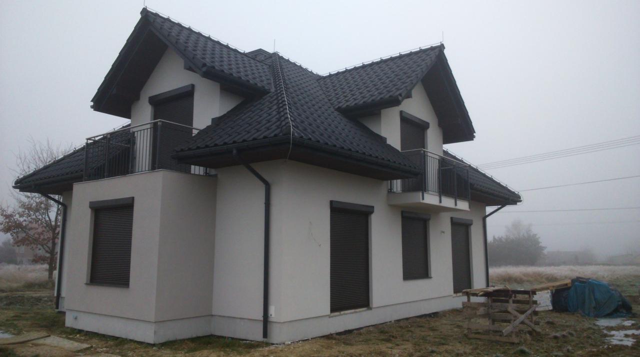 Dwupiętrowy dom z czarnym dachem i roletami, widziany z boku w pochmurny dzień. Balkony z czarną barierką. Na trawie widoczne materiały budowlane.