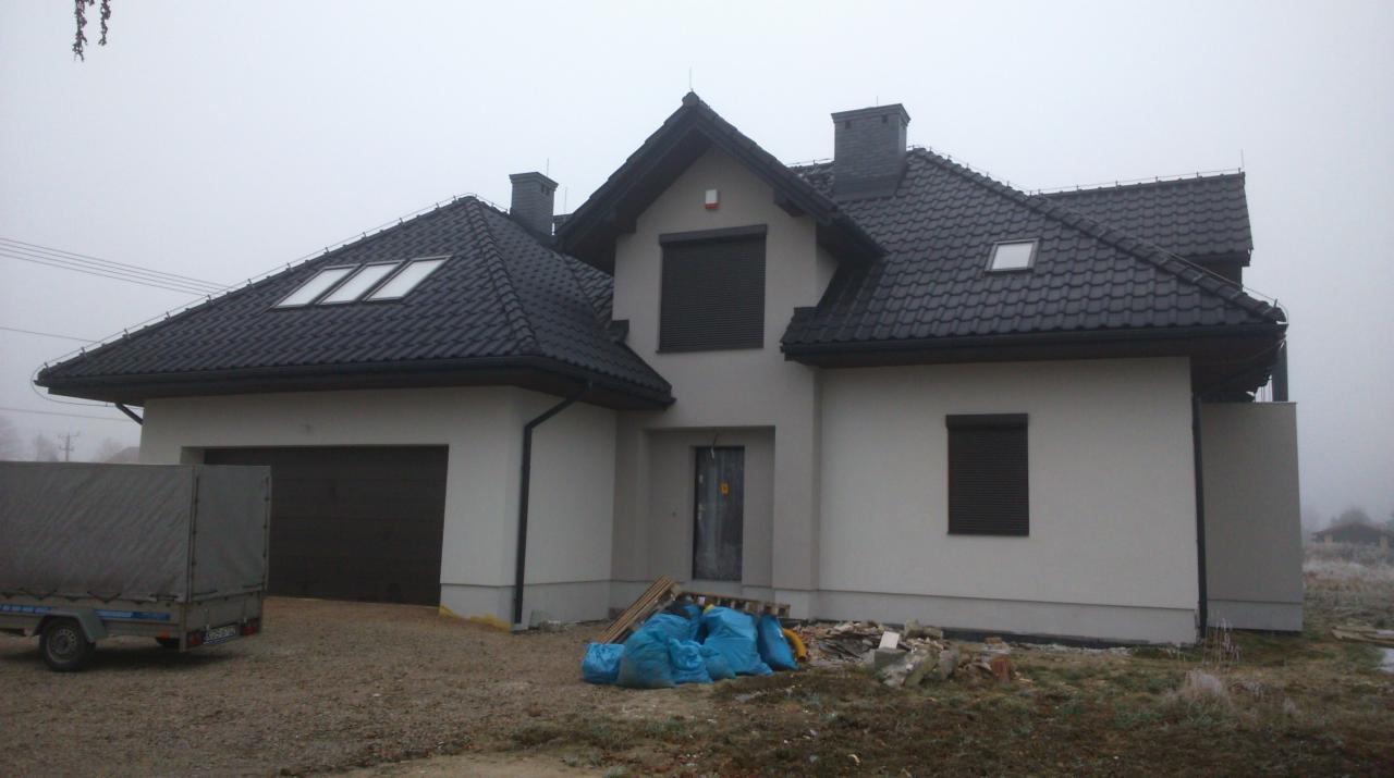 Nowoczesny dom z ciemną dachówką i jasną elewacją w pochmurny dzień. Widoczny garaż z bramą, okna z roletami i niebieskie worki przed wejściem.