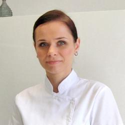 Portret kobiety w białym uniformie, prawdopodobnie kosmetyczki lub lekarki, z delikatnym makijażem i upiętymi włosami, na jasnym tle.