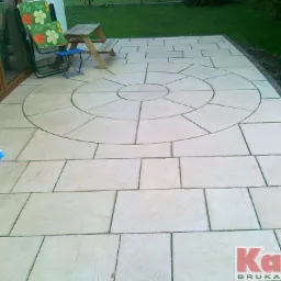 Jasna kostka brukowa ułożona w okrągły wzór na tarasie, z krzesłem i stolikiem w tle. Widoczne logo firmy brukarskiej w prawym dolnym rogu.