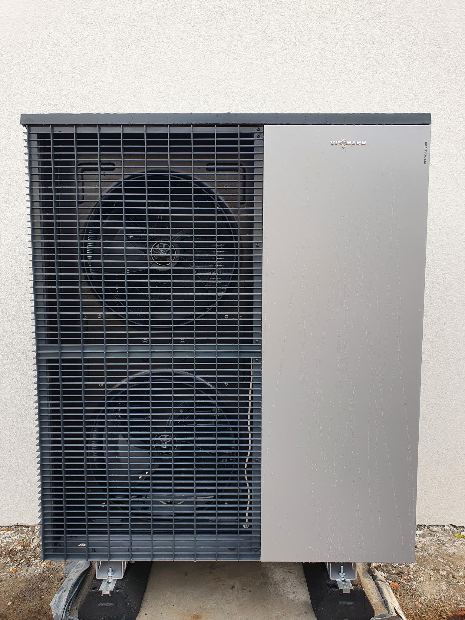Szara pompa ciepła Viessmann Vitocal 200, stojąca na zewnątrz budynku, z widocznymi dwoma wentylatorami za metalową kratą i kroplami wody na obudowie.