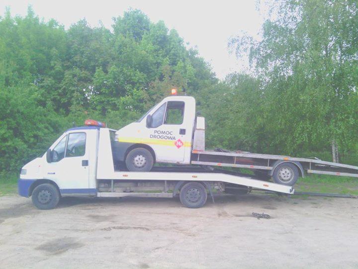 Laweta z pojazdem 'Pomoc Drogowa' na platformie, na tle zieleni. Widoczna sygnalizacja świetlna na obu pojazdach. Ujęcie z poziomu gruntu.