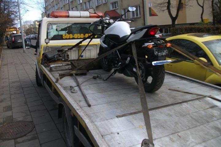 Biały motocykl zabezpieczony pasami na platformie lawety z pomarańczowym światłem ostrzegawczym. W tle budynek mieszkalny i żółty samochód osobowy.