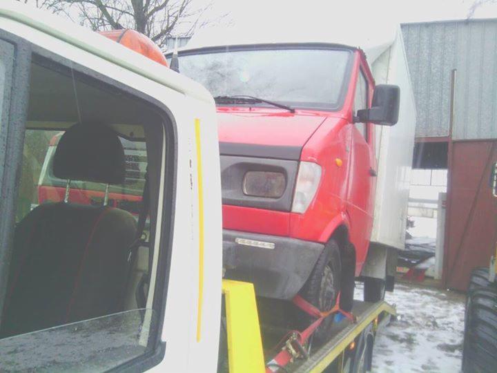 Czerwony samochód dostawczy na żółtej lawecie, zabezpieczony pasami transportowymi, częściowo widoczny biały pojazd z lewej strony kadru.