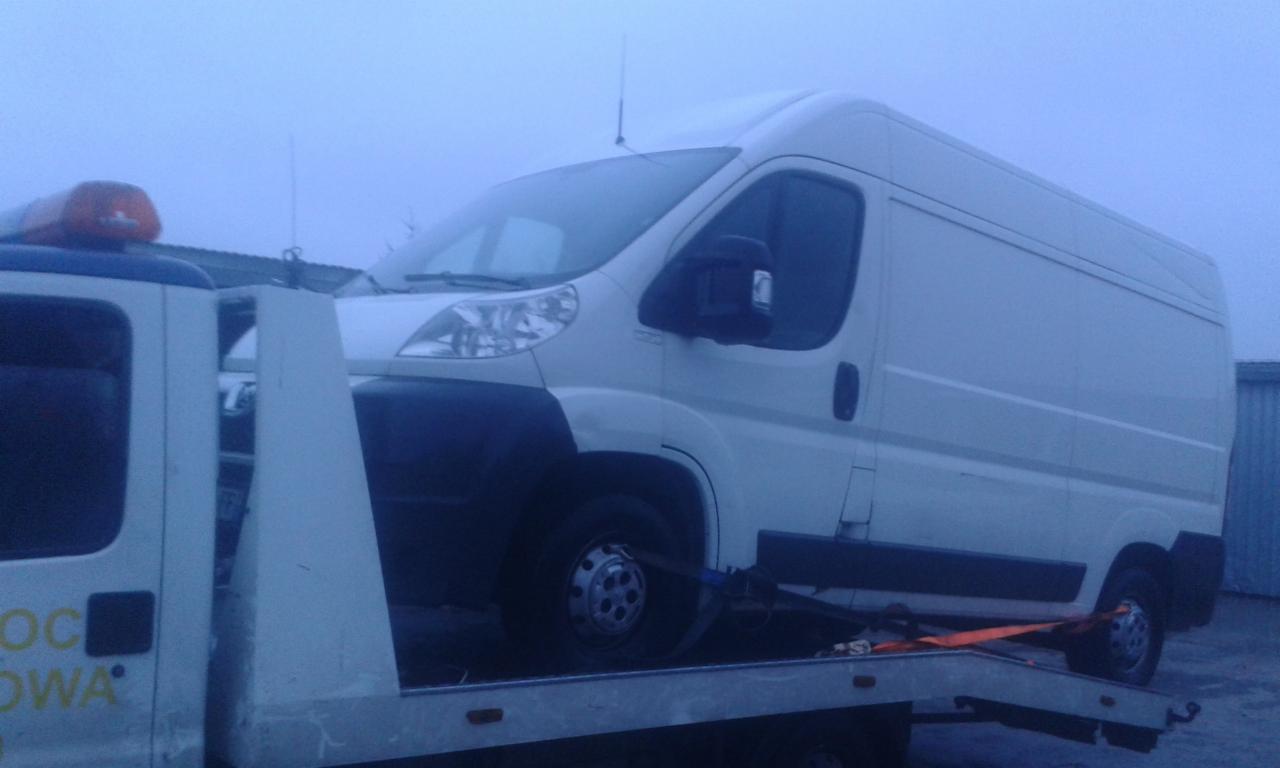 Biały bus na lawecie w pochmurny dzień. Pomarańczowe pasy zabezpieczają pojazd. Widoczna lampa ostrzegawcza na lawecie.