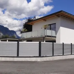 Nowoczesna posesja z aluminiowym ogrodzeniem panelowym w kolorze srebrnym i czarnym, na tle białego domu z balkonem i górskim krajobrazem w tle.