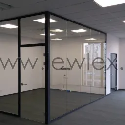 Szklana ścianka działowa z czarnymi profilami w biurze, z widocznym logo firmy na szkle i oknami z widokiem na budynek mieszkalny.
