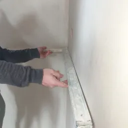 Mężczyzna w szarej bluzie przykłada poziomicę w kształcie litery L do świeżo otynkowanej ściany w rogu pomieszczenia, widoczne ślady gładzi na ścianie i narzędziu.