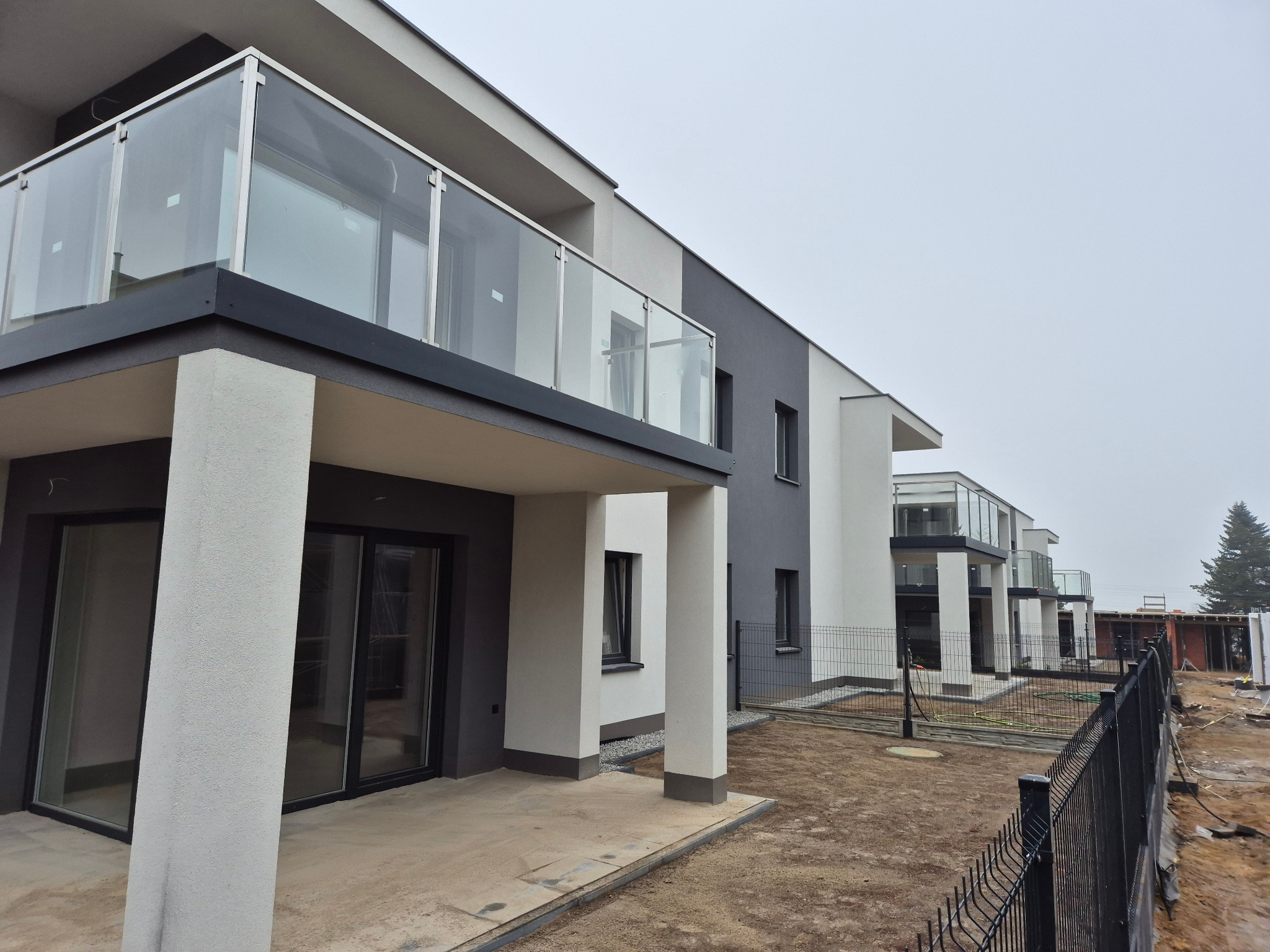 Nowoczesny budynek wielorodzinny z balkonami o szklanych balustradach, pomalowany w odcieniach szarości i bieli, widoczny z zewnątrz na etapie zagospodarowywania terenu.