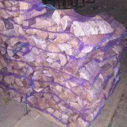 Stos worków z pociętym drewnem kominkowym, ułożonych na palecie, widoczny wózek transportowy w tle.