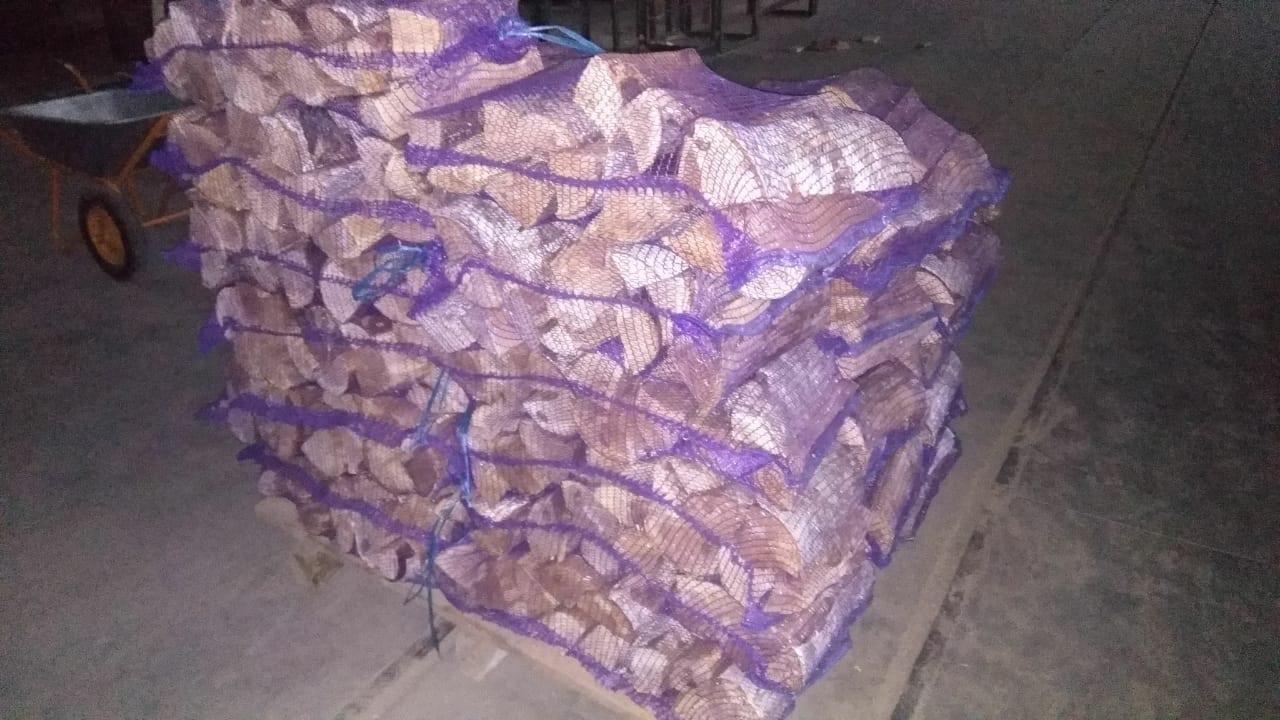 Stos worków z pociętym drewnem kominkowym, ułożonych na palecie, widoczny wózek transportowy w tle.