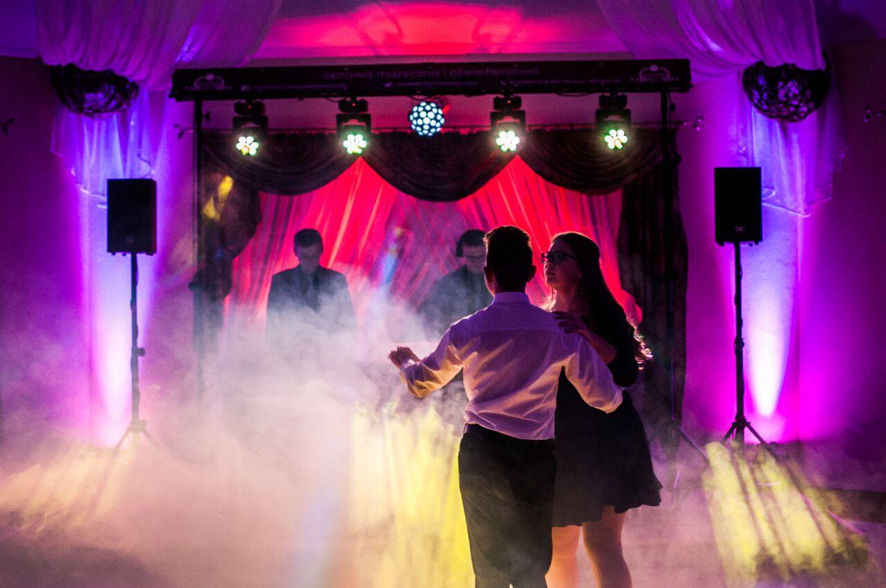 Para tańczy na parkiecie w oparach dymu, w tle scena z czerwonymi zasłonami, reflektorami i kolumnami głośnikowymi