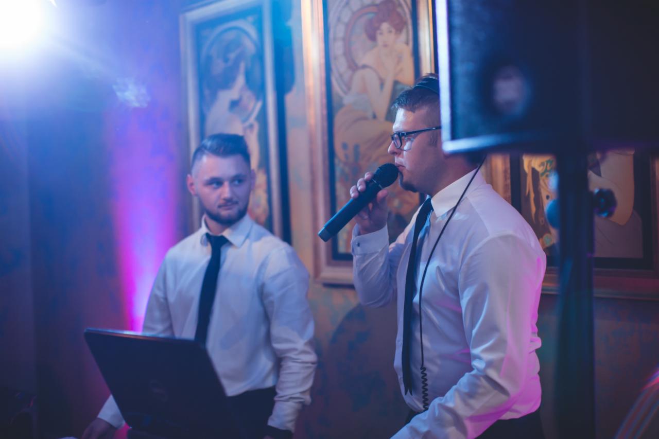 Mężczyzna w okularach śpiewa do mikrofonu na tle obrazów w ramkach, drugi mężczyzna obsługuje laptopa. Oświetlenie sceniczne.