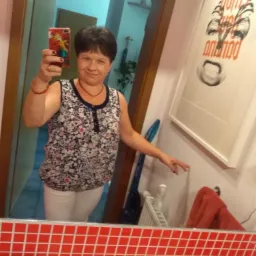 Odbicie w lustrze kobiety robiącej selfie w łazience z czerwonymi kafelkami, widoczny odkurzacz pionowy i plakat na ścianie.