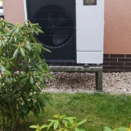 Zewnętrzna jednostka pompy ciepła Mitsubishi Zubadan R32 zamontowana na metalowym stelażu obok budynku z elewacją w kolorze brzoskwini, widoczna zza krzewu rododendrona.