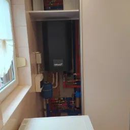 Zamontowany, szary piec gazowy Termo+ z niebieskim naczyniem przeponowym, widoczne rury instalacji w kolorach czerwonym i niebieskim, zamontowane w rogu pomieszczenia z jasnymi płytkami.