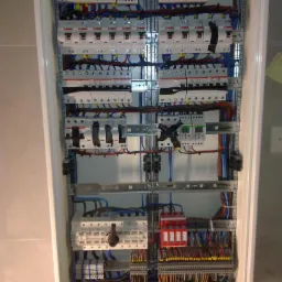 Rozbudowana skrzynka elektryczna z licznymi bezpiecznikami, przekaźnikami i okablowaniem, zamontowana w ścianie wyłożonej płytkami ceramicznymi.