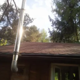 Zewnętrzny komin ze stali nierdzewnej, zamontowany na drewnianym budynku z dachem pokrytym gontem bitumicznym, widoczne drzewa w tle.