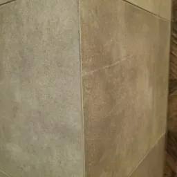 Szczegółowe ujęcie narożnika wykończonego płytkami w odcieniach beżu i szarości, z widocznymi fugami i delikatną fakturą płytek, po prawej stronie fragment ściany wykończonej drewnem.