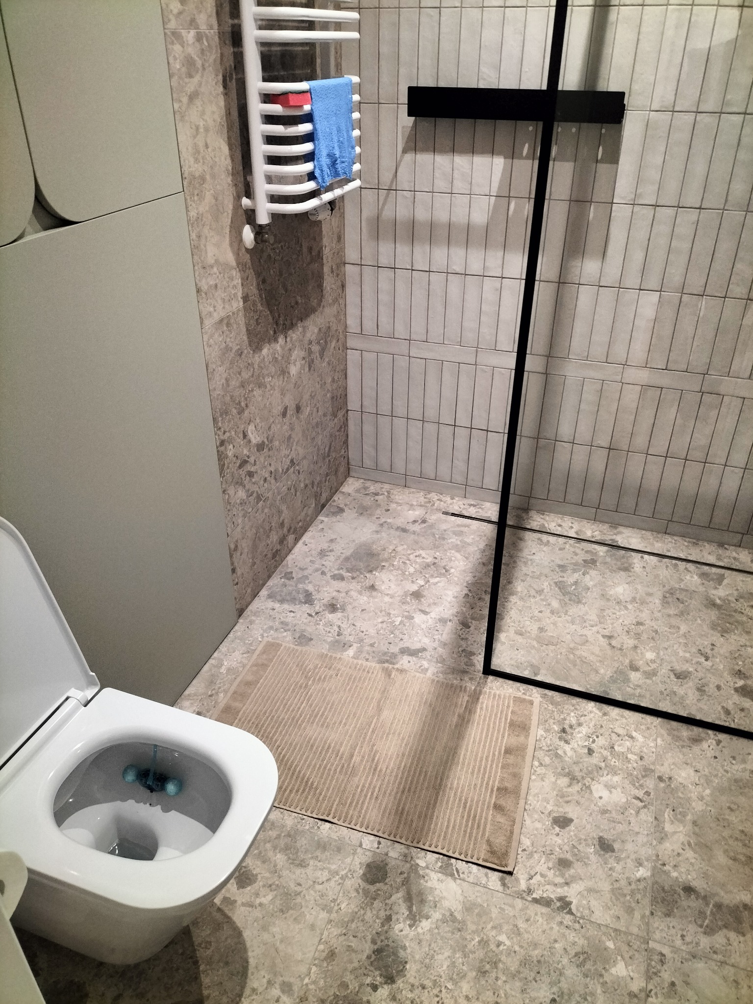 Nowoczesna łazienka z szarymi płytkami imitującymi marmur, kabiną prysznicową z czarną ramą, toaletą wiszącą z otwartą deską, grzejnikiem drabinkowym z niebieskim ręcznikiem i beżowym dywanikiem.