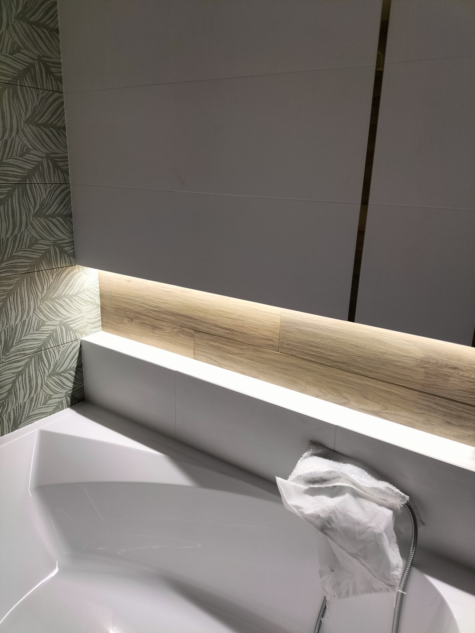 Nowoczesna łazienka: biała wanna z ukrytą listwą LED imitującą drewno, ściana z płytek w liście i szara płytka z cienkim złotym paskiem, zabezpieczona słuchawka prysznicowa.
