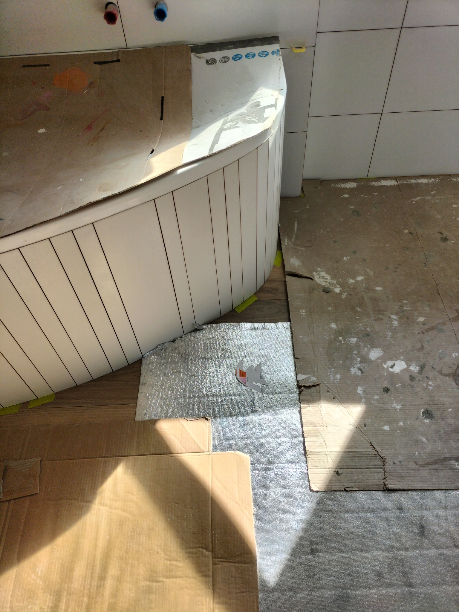 Przygotowanie do montażu obudowy wanny z płytek ceramicznych, zabezpieczone podłoże kartonem i folią aluminiową, widoczne fragmenty płytek na ścianie.