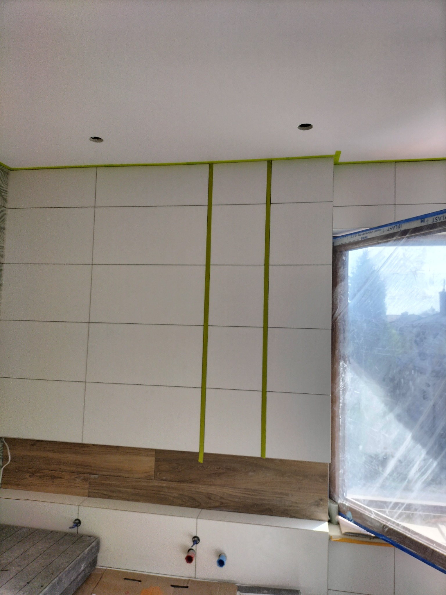 Ściana w trakcie układania białych, prostokątnych płytek z drewnopodobnym dekorem w dolnej części, zabezpieczona taśmą malarską, widok z Piaseczna.