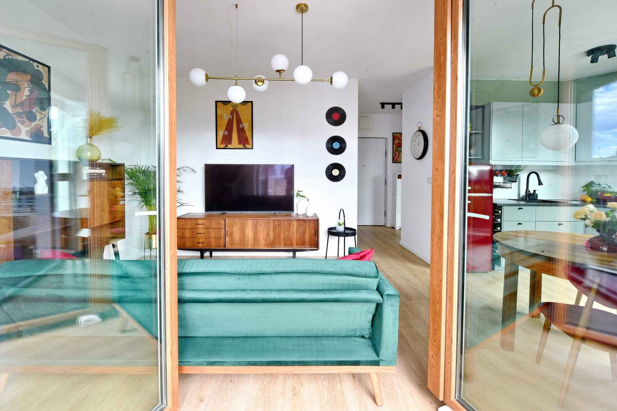 Nowoczesne wnętrze mieszkania z otwartą kuchnią, widok z salonu przez szklane drzwi balkonowe, turkusowa sofa, drewniane meble w stylu retro, gramofonowe płyty na ścianie.