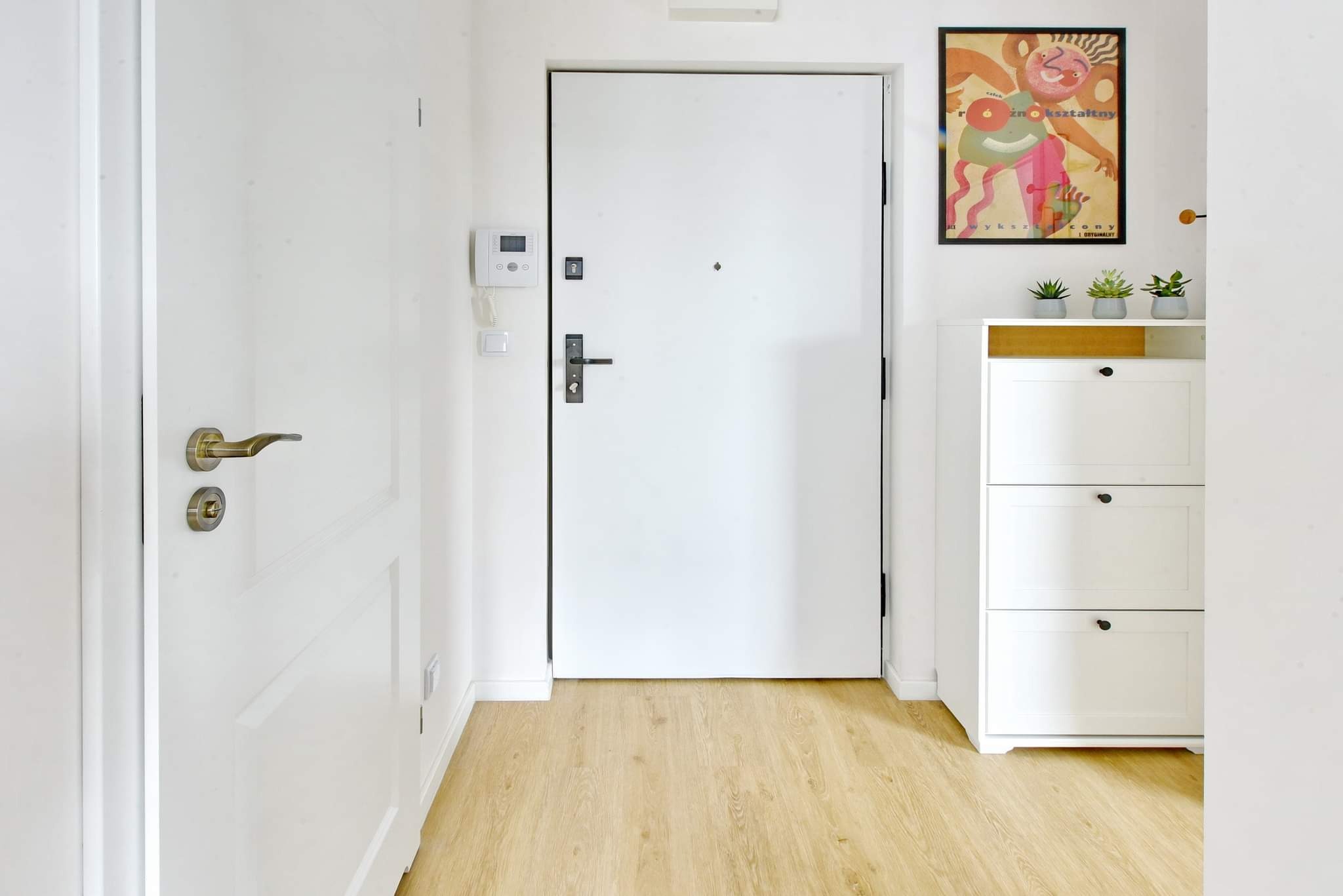 Jasne wnętrze przedpokoju z białymi drzwiami wejściowymi, panelami podłogowymi w odcieniach drewna i białymi meblami na buty, nad którymi wisi kolorowy plakat.