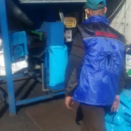 Pracownik w niebieskiej kamizelce z napisem 'RE-TRANS' i dużym motywem pająka na plecach, stoi obok niebieskiej maszyny pakującej i zapełnionych worków z materiałem.