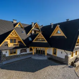 Nowoczesny dom w stylu góralskim z ciemnym dachem i jasną elewacją, wykończony kamieniem i drewnem, z widocznym logo na elewacji, na tle błękitnego nieba.