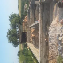 Wznoszenie murów fundamentowych z bloczków betonowych na budowie. Widoczne narzędzia i materiały budowlane. W tle zieleń i niebo.