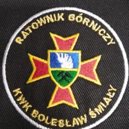 Czarna naszywka z haftowanym, kolorowym symbolem górniczym: krzyż z motywem dłoni, narzędzi górniczych i napisem 'Ratownik Górniczy KWK Bolesław Śmiały'.