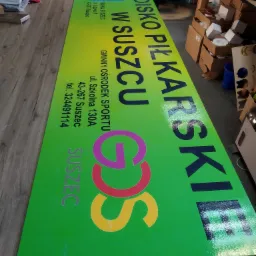 Duży, zielony baner reklamowy z napisem 'Boisko Piłkarskie w Suszcu' i logo GOS, leżący na stole w pomieszczeniu biurowym.