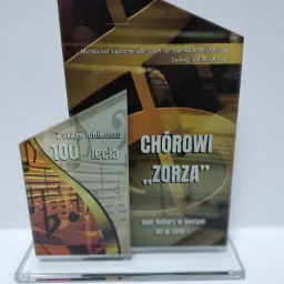 Szklana statuetka z okazji 100-lecia chóru 'Zorza', zawierająca cytat Ludwiga van Beethovena o muzyce, umieszczona na przezroczystej podstawie.